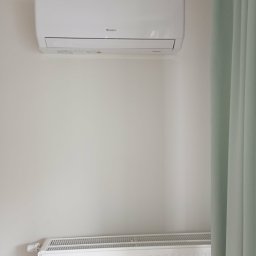 WOJ-TECH Wojciech Świderski - Biały klimatyzator marki Gree zamontowany na ścianie nad białym grzejnikiem, obok zwisającej zielonej zasłony. Minimalistyczny design wnętrza.