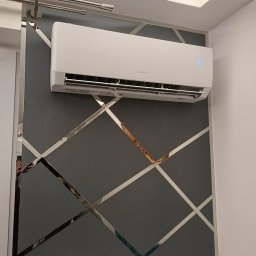 WOJ-TECH Wojciech Świderski - Biała klimatyzacja Gree na szarej ścianie z geometrycznym wzorem z lustrzanych pasków. Widoczny wyświetlacz temperatury. Nowoczesny design.