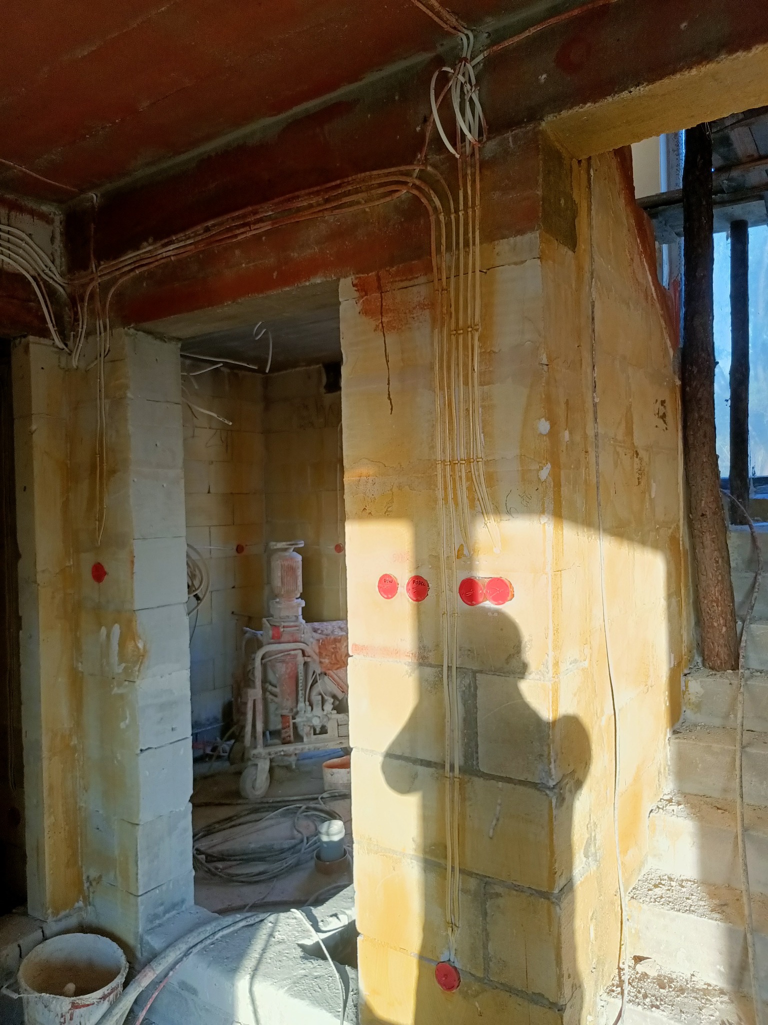 Surowy stan budynku w trakcie prac instalacyjnych: widoczne przewody elektryczne prowadzone po ścianach z czerwonymi puszkami, sprzęt budowlany w tle, cień osoby na ścianie.