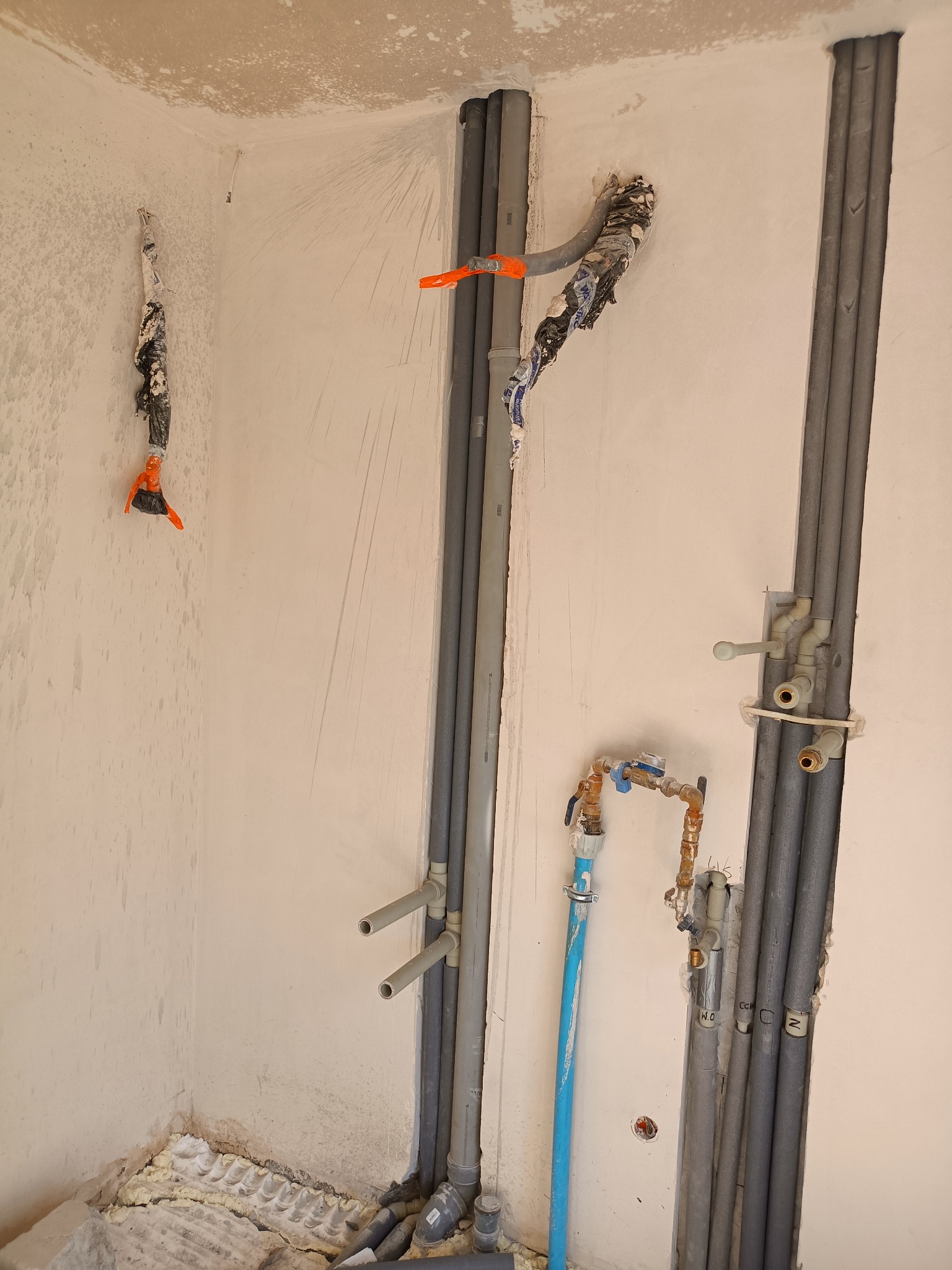 Szare rury instalacji wodnej i kanalizacyjnej w bruzdach ściany, z zaworami i przyłączami, w surowym stanie przed wykończeniem. Widoczne zabezpieczenia rur.