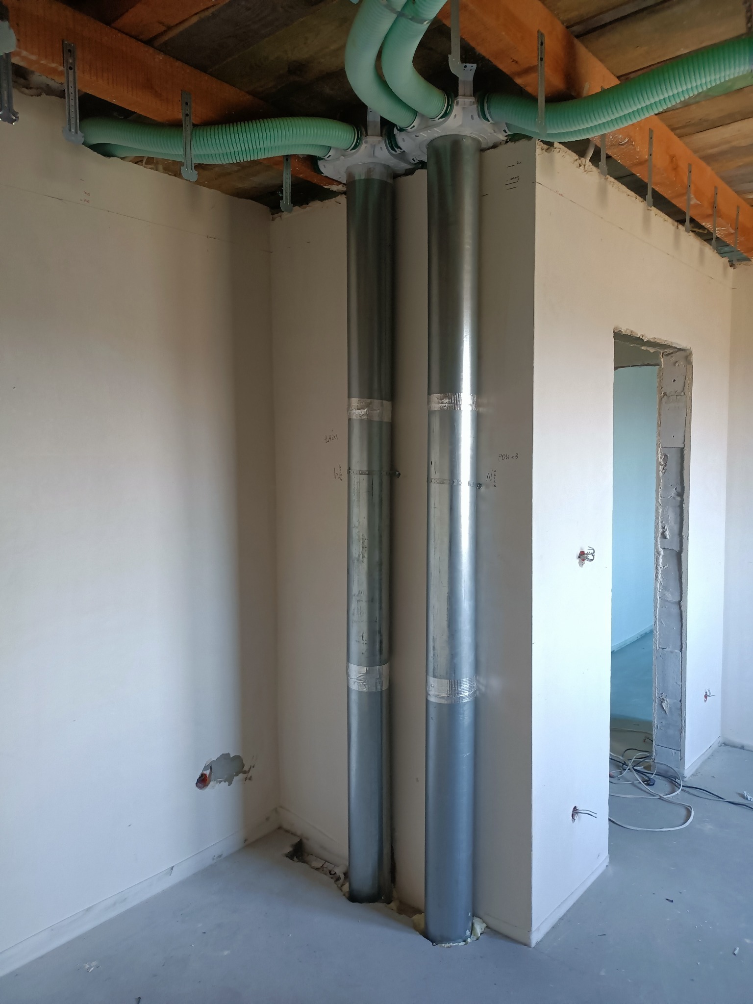 Dwie pionowe, metalowe rury wentylacyjne z zielonymi, elastycznymi przewodami pod sufitem w surowym wnętrzu. Widoczne ślady po instalacji i niedokończone ściany.