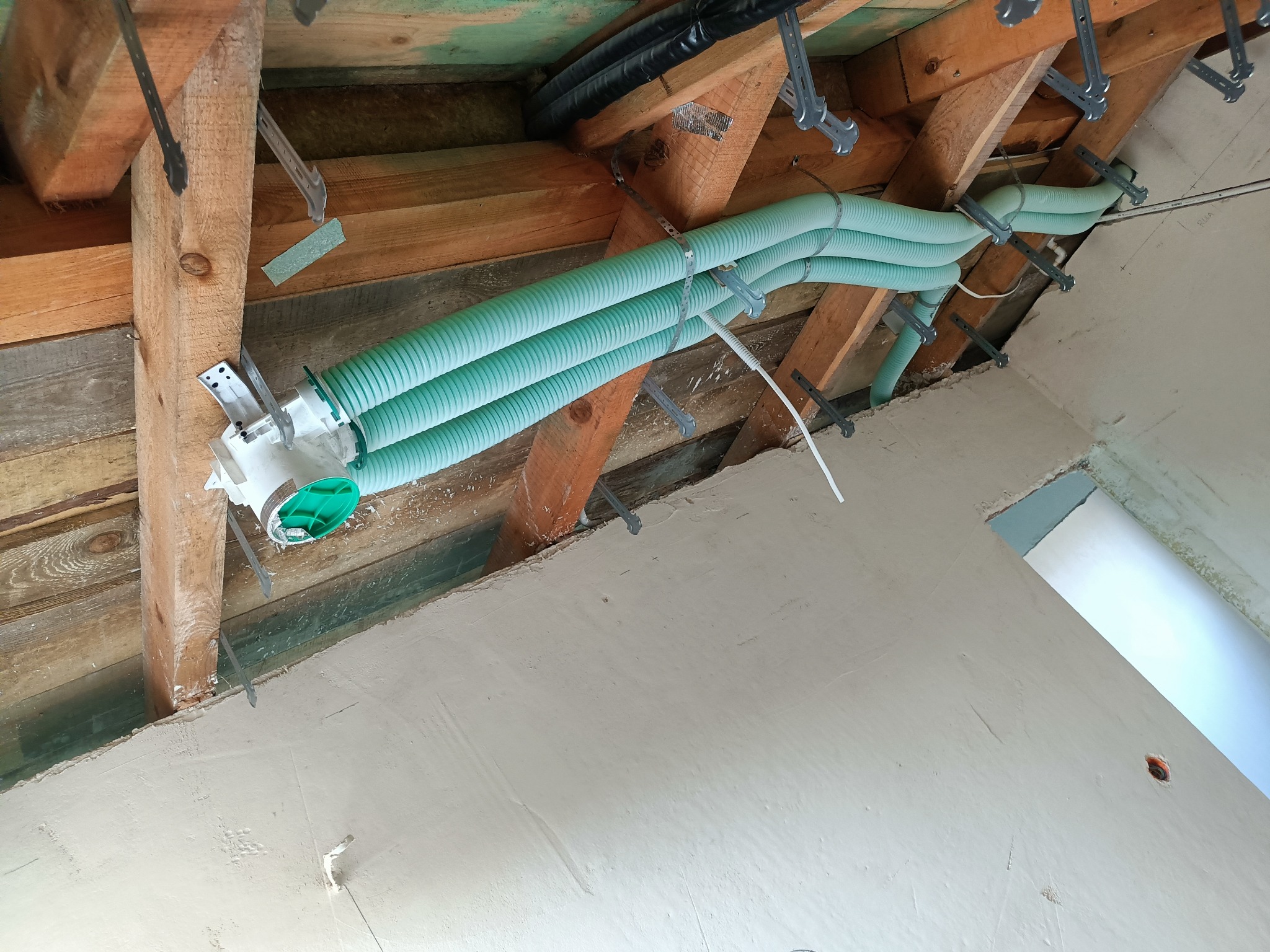 Zielone rury wentylacyjne zamocowane do drewnianej konstrukcji dachu na poddaszu, widoczne elementy instalacji i fragment otynkowanej ściany.