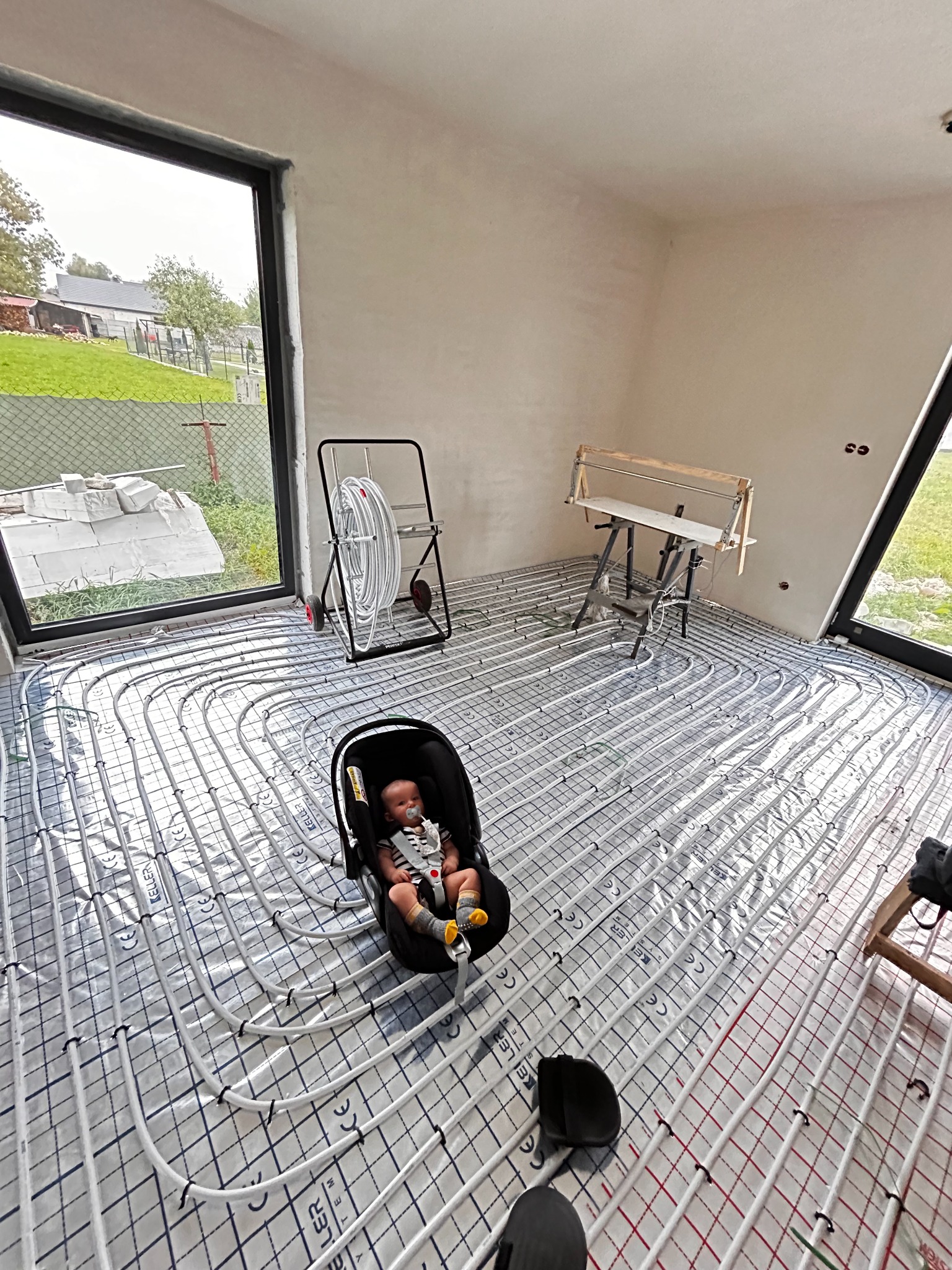 Białe rury ogrzewania podłogowego ułożone na folii z siatką, w tle okno i zwinięte rury na stojaku. W pokoju jest dziecko w foteliku samochodowym.