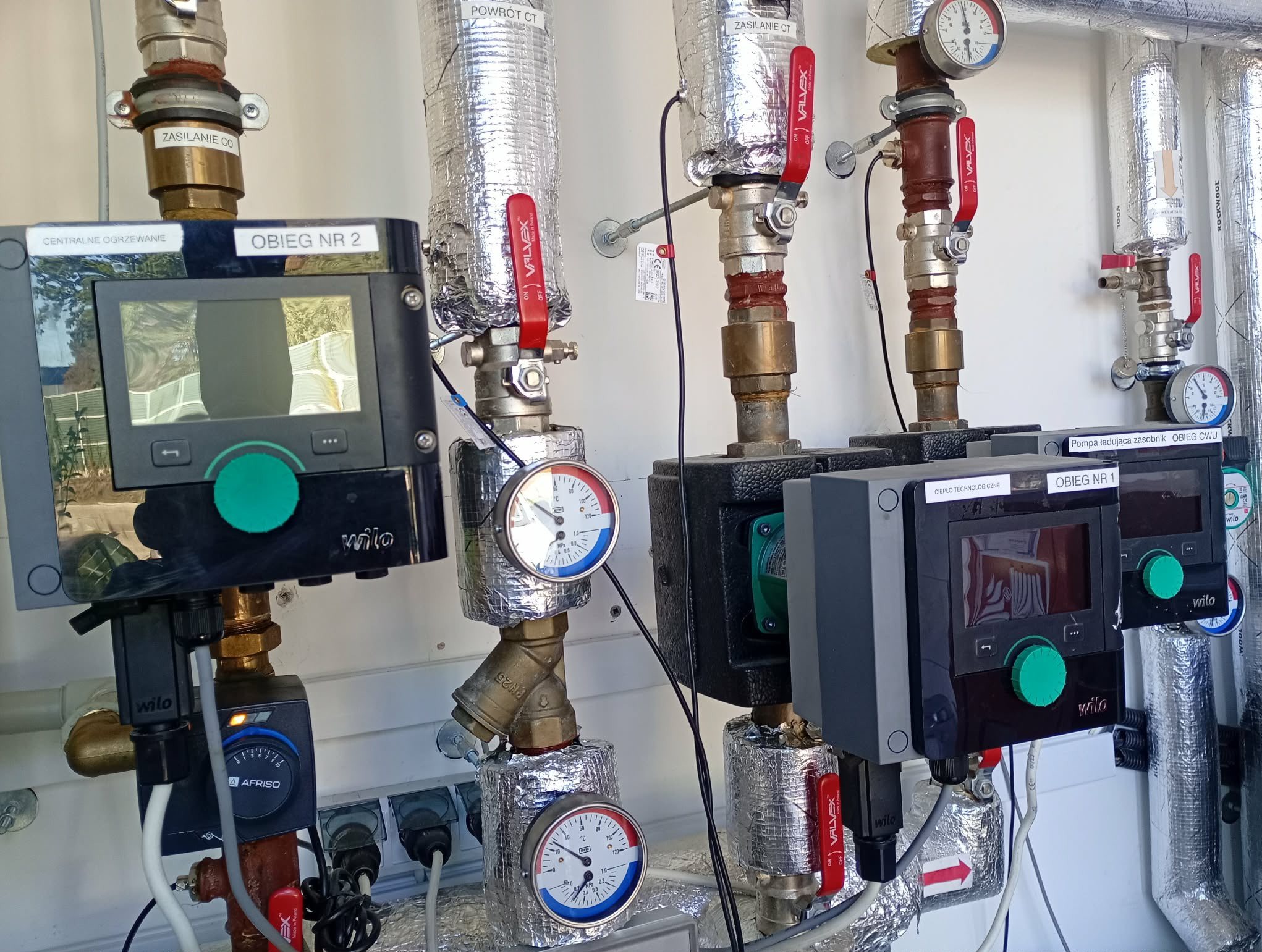 Węzeł cieplny z dwoma obiegami, widoczne pompy obiegowe Wilo, zawory Valvex, manometry, izolowane rury i sterowniki z wyświetlaczami. Elementy instalacji centralnego ogrzewania.