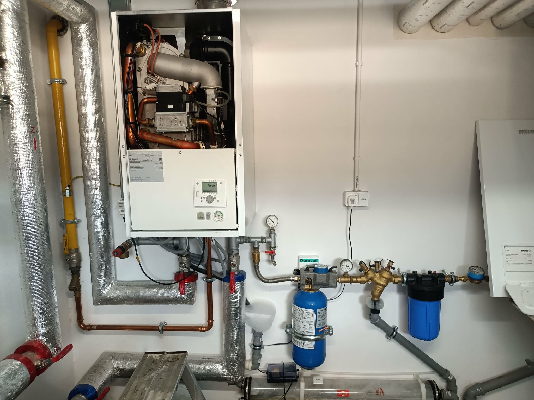 Nowoczesny piec gazowy z filtrami i rurami w kotłowni w Kosowie Lackim. Widoczne elementy instalacji grzewczej i wodnej. Białe ściany, schludne wykonanie.
