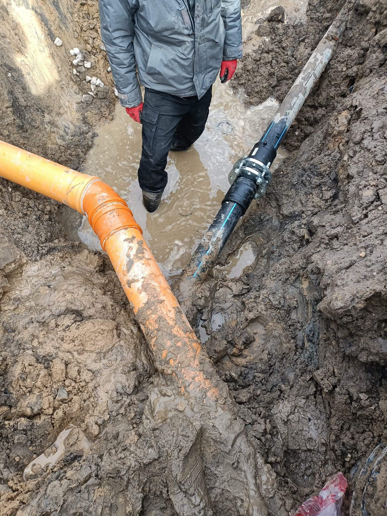 Połączenie rur w wykopie wypełnionym wodą. Widoczne pomarańczowe rury kanalizacyjne i czarna rura z metalowym łącznikiem. Osoba w roboczym ubraniu stoi w wodzie.