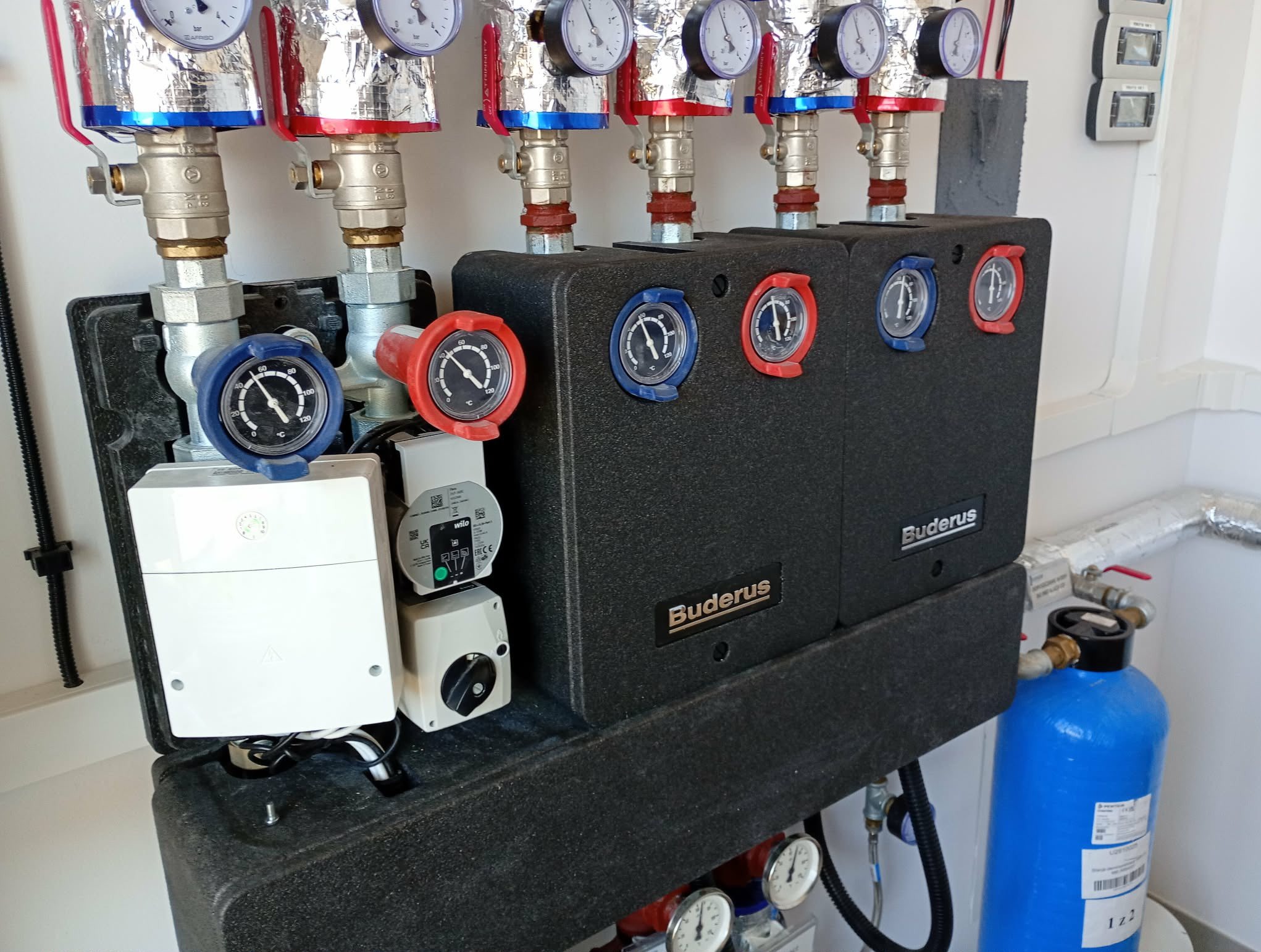 Rozdzielacz ogrzewania podłogowego Buderus z manometrami i pompami obiegowymi, widoczne rury z izolacją termiczną i filtr wody. Element instalacji grzewczej w kotłowni.