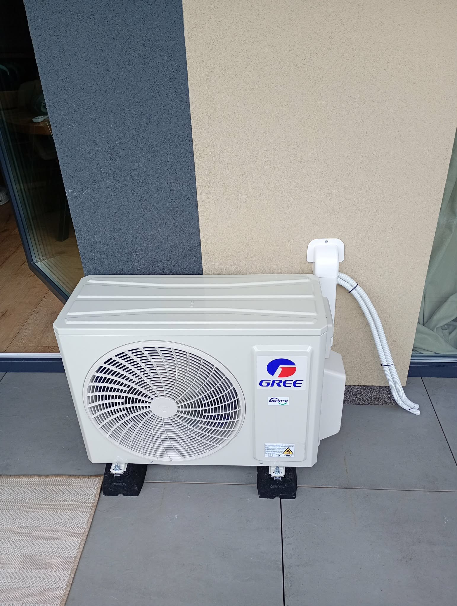 Biała jednostka zewnętrzna klimatyzacji Gree zamontowana na balkonie, z widocznymi przewodami i elementami mocującymi. Tło stanowi elewacja budynku w dwóch odcieniach.
