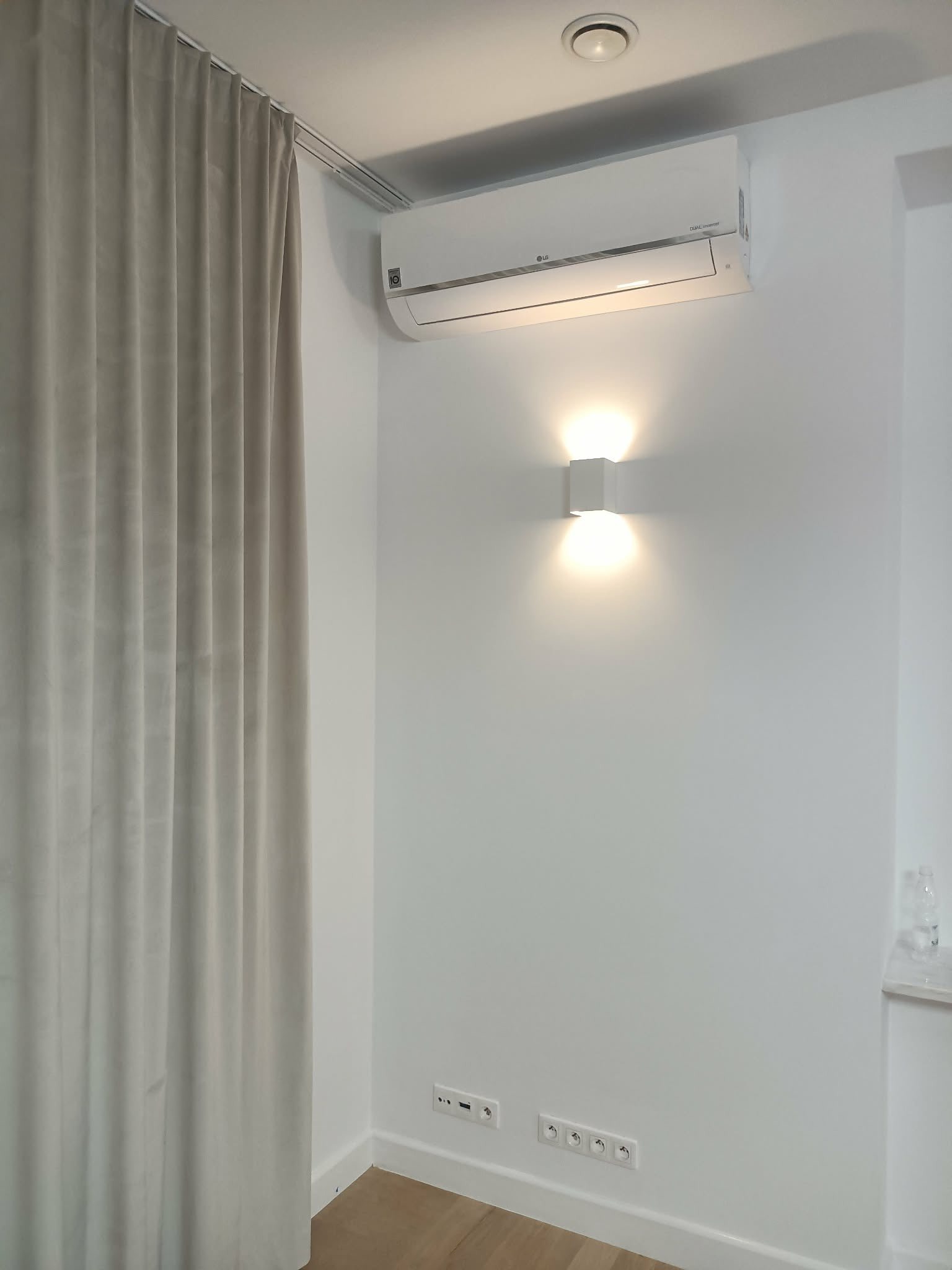 Biały klimatyzator LG na ścianie nad kwadratową lampą ścienną, obok zasłony. Widoczne gniazdka elektryczne. Minimalistyczne wnętrze.