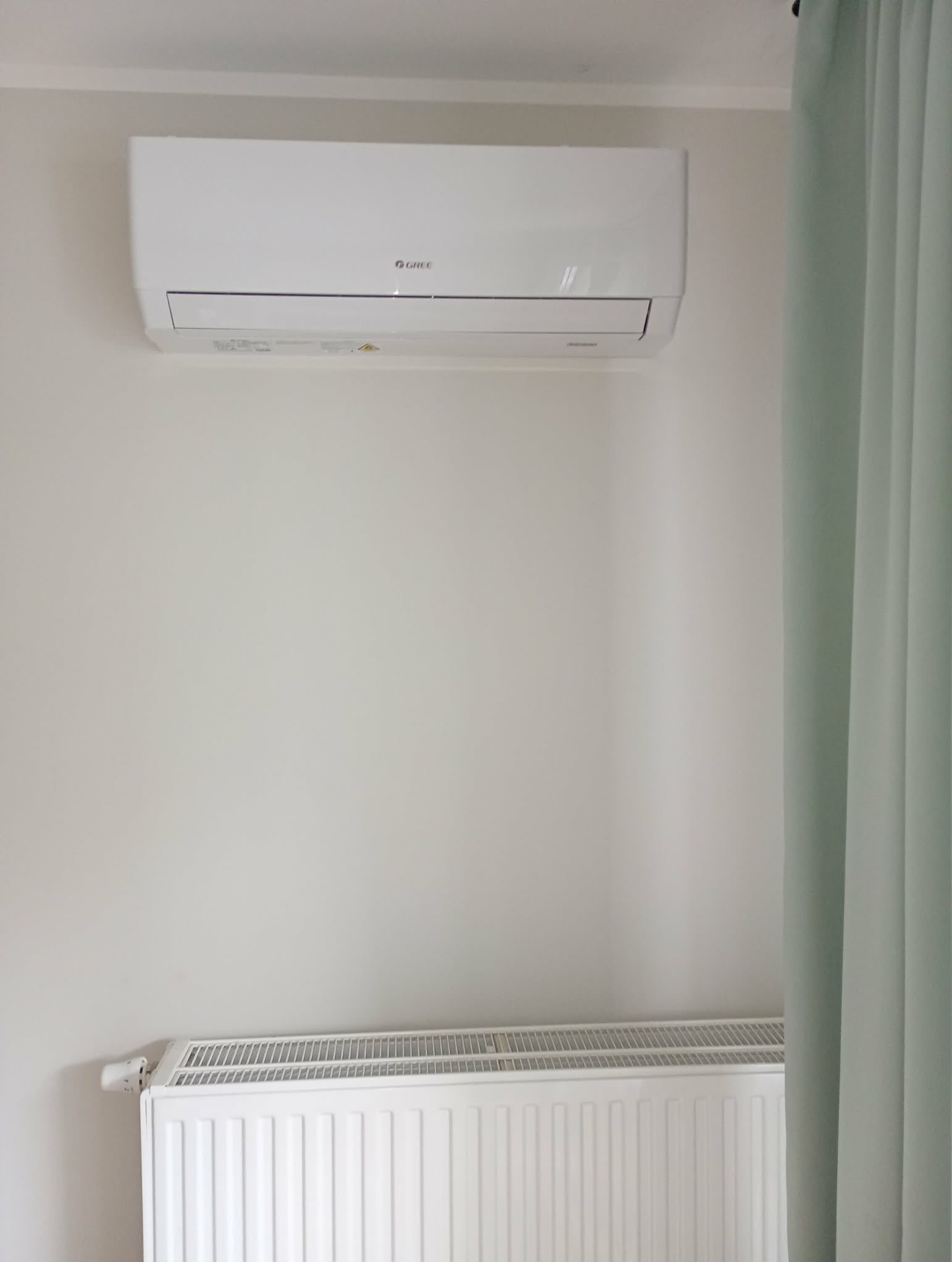 Biały klimatyzator marki Gree zamontowany na ścianie nad białym grzejnikiem, obok zwisającej zielonej zasłony. Minimalistyczny design wnętrza.