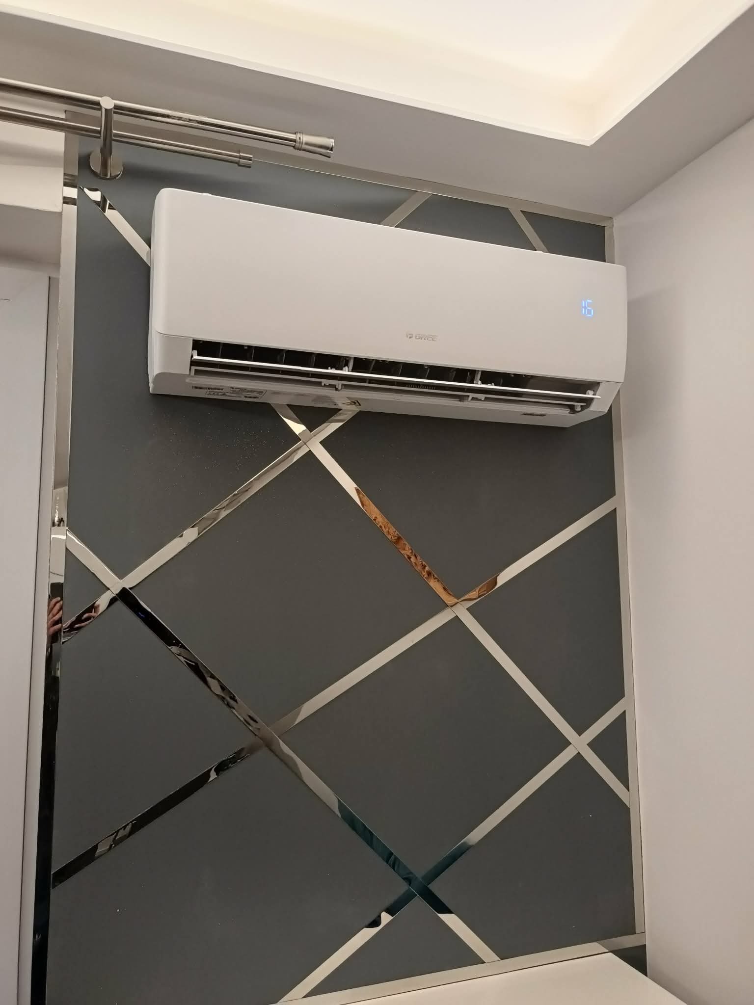 Biała klimatyzacja Gree na szarej ścianie z geometrycznym wzorem z lustrzanych pasków. Widoczny wyświetlacz temperatury. Nowoczesny design.