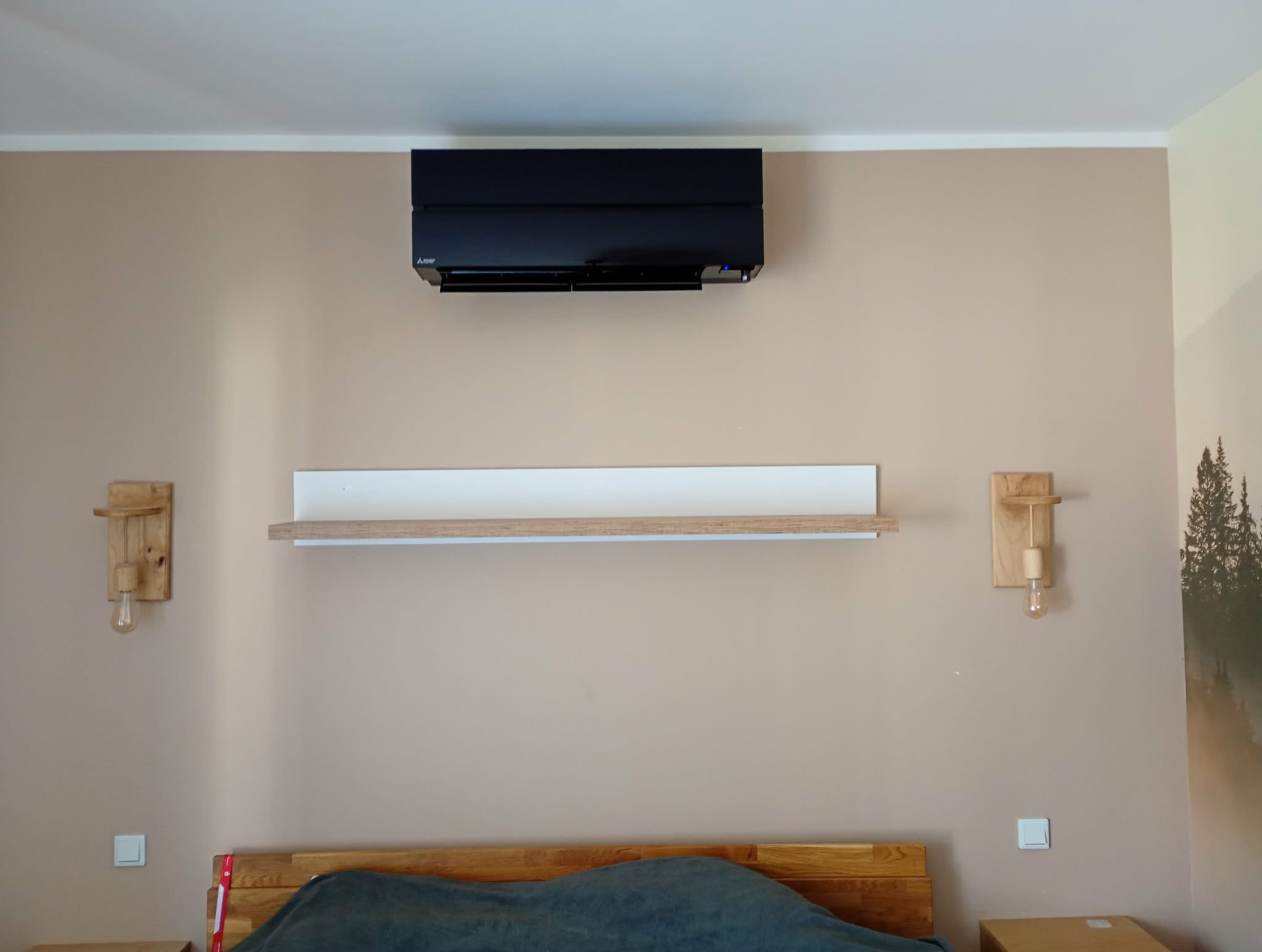 Czarny klimatyzator Mitsubishi Electric zamontowany nad półką w sypialni, z drewnianymi lampkami po bokach i łóżkiem w dolnej części kadru. Beżowe ściany.
