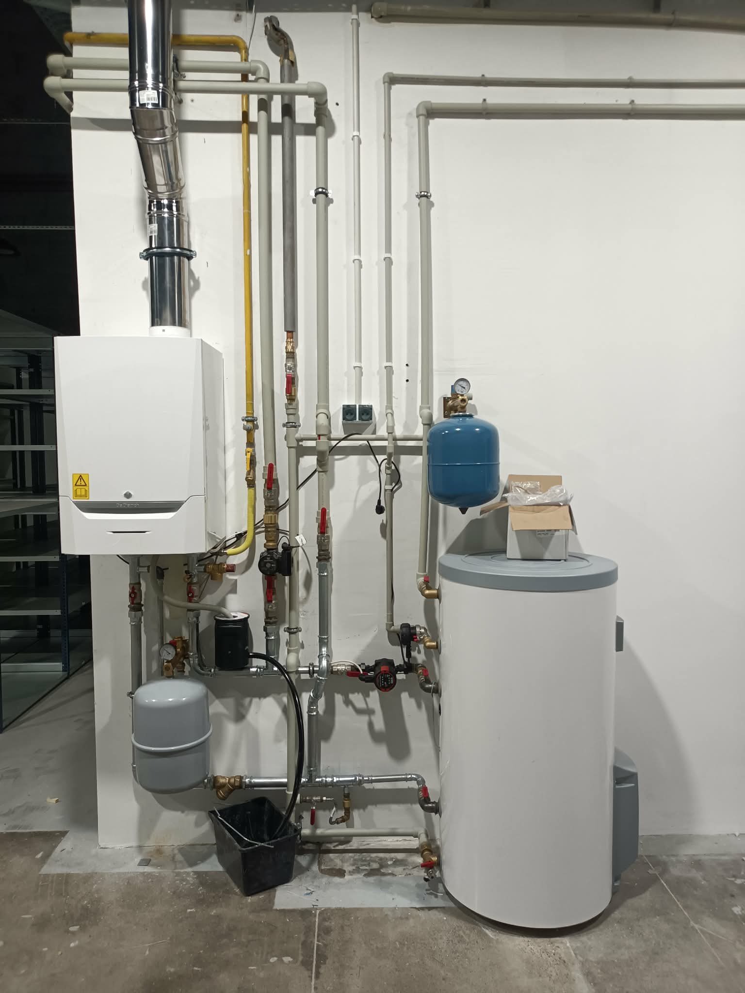 Biały piec gazowy kondensacyjny z podłączeniami, zbiornik wody i rury na tle białej ściany. Widoczny manometr i zawory. Na podłodze wiadro i karton.