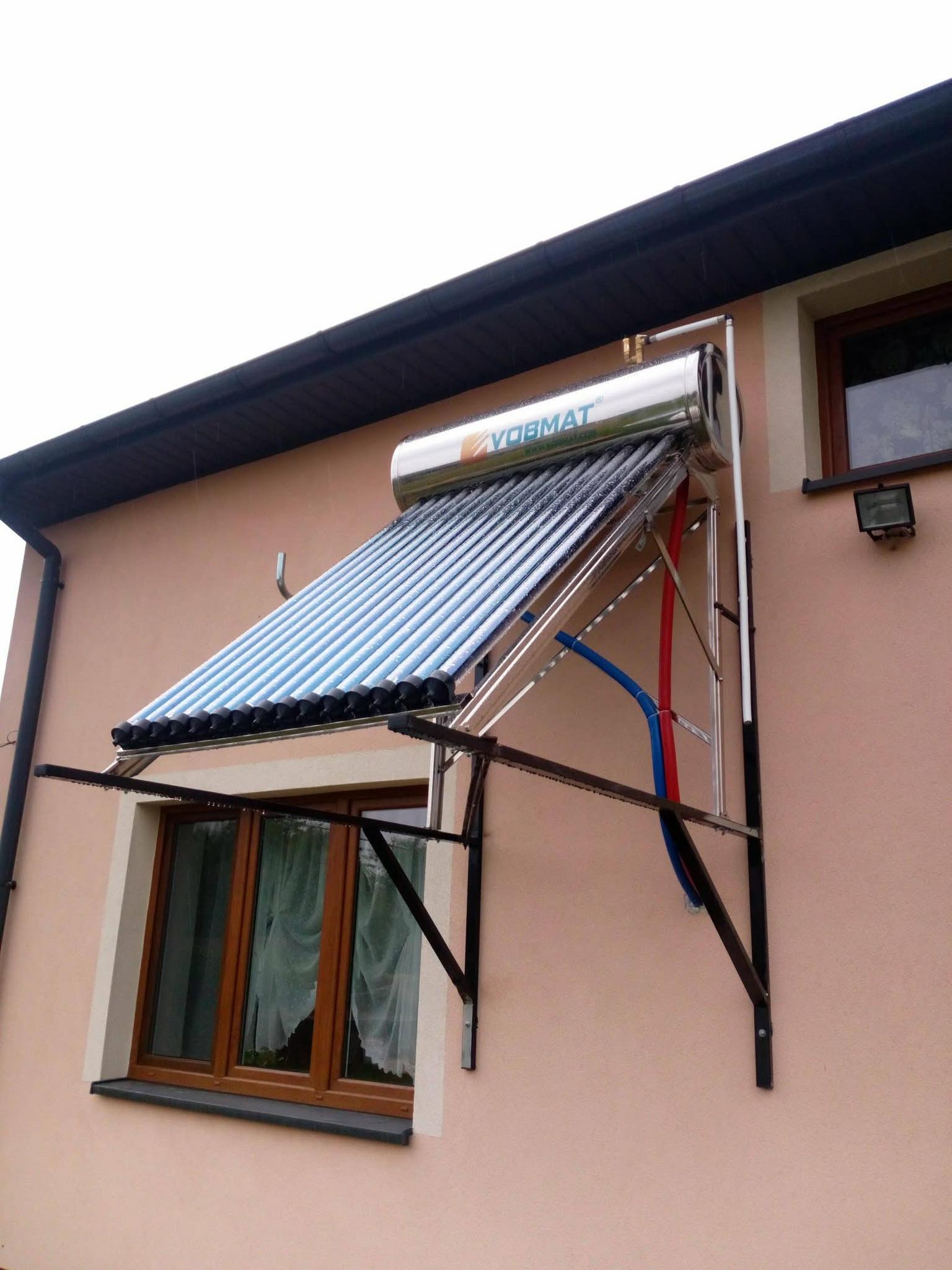 Kolektor słoneczny VOBMAT zamontowany nad oknem na elewacji domu. Widoczne rury doprowadzające wodę (czerwona i niebieska).