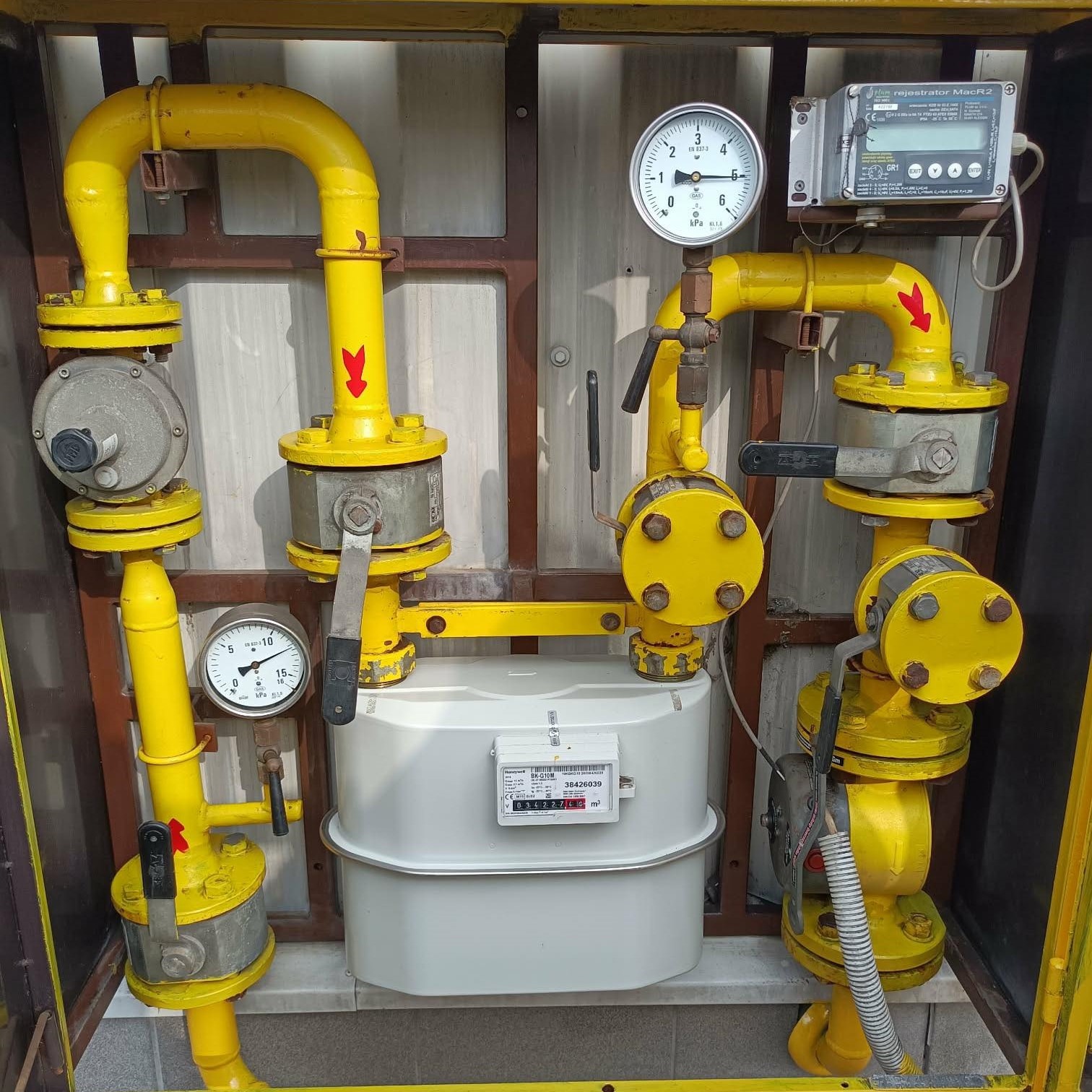 Żółta instalacja gazowa z manometrami i licznikiem w metalowej obudowie. Widoczne zawory, rury i elektroniczny rejestrator. Elementy instalacji pomalowane na żółto.