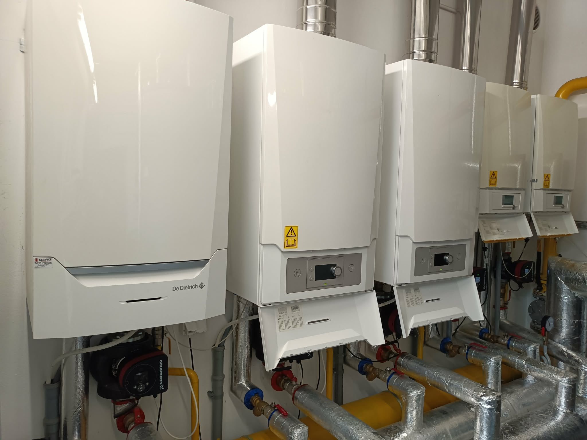 Rząd białych, nowoczesnych pieców gazowych De Dietrich z widocznymi rurami instalacji grzewczej, zamontowanych na ścianie w pomieszczeniu technicznym.