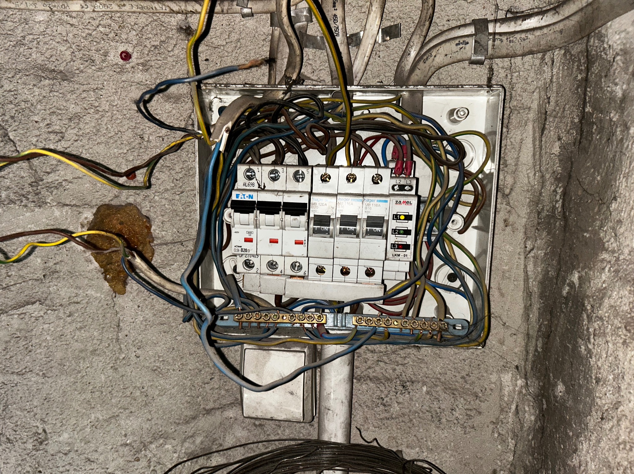 Otwarta skrzynka elektryczna z bezpiecznikami Eaton i Hager, widoczne przewody w kolorach niebieskim, żółtym i brązowym, zamontowana na szarej, surowej ścianie.