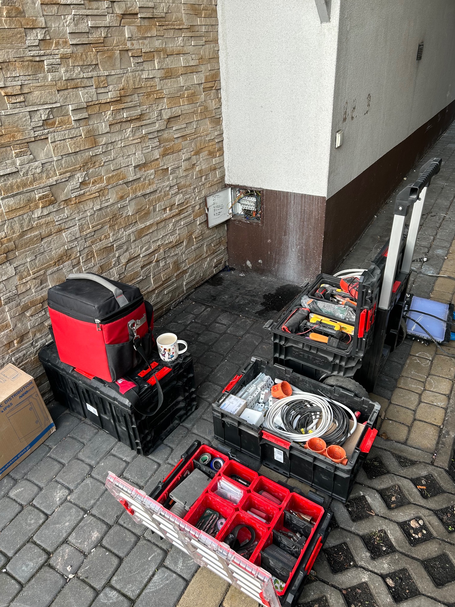 Otwarty rozdzielacz elektryczny na ścianie budynku, obok skrzynki z narzędziami i akcesoriami elektrycznymi, kubek z kawą, drabina. Praca w toku.