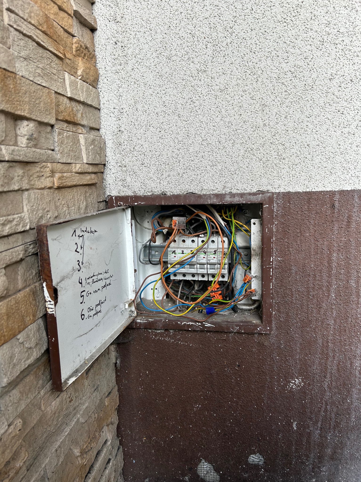 Otwarta skrzynka elektryczna z widocznymi przewodami i listą kontrolną na drzwiczkach, zamontowana na ścianie z elementami dekoracyjnymi.