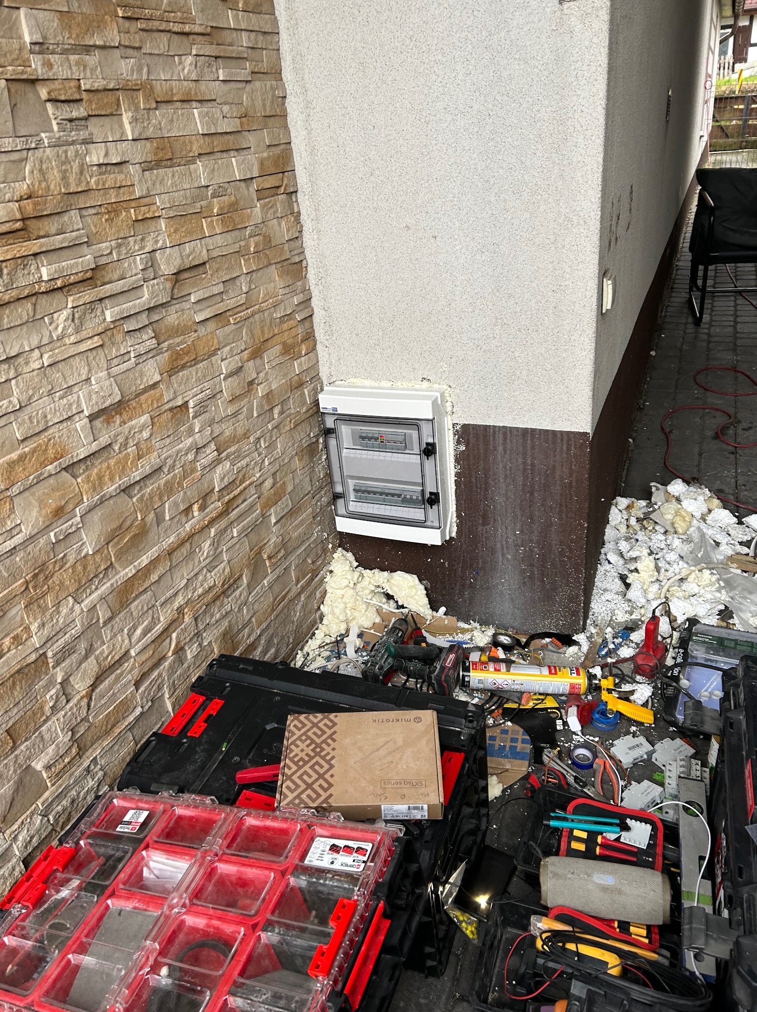 Zamontowana skrzynka elektryczna na elewacji budynku, widoczne narzędzia i materiały instalacyjne. Prace w toku, pianka montażowa wokół skrzynki.