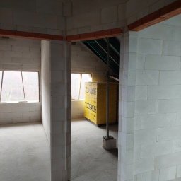 MaxBud Maksym Maruda - Wnętrze domu w trakcie budowy z białych bloczków Ytong, widoczne okna i drewniane belki stropowe. Na podłodze wylewka, w tle żółte opakowania bloczków.