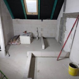 MaxBud Maksym Maruda - Wnętrze poddasza w trakcie budowy ścian z białych bloczków, widoczne narzędzia, poziomica i wiadro. Ukończona ściana z oknem dachowym. Etap prac murarskich.