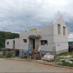 MaxBud Maksym Maruda - Budowa domu z białych bloczków, widoczne ściany w stanie surowym otwarte, teren budowy zabezpieczony siatką, w tle zieleń i pochmurne niebo.