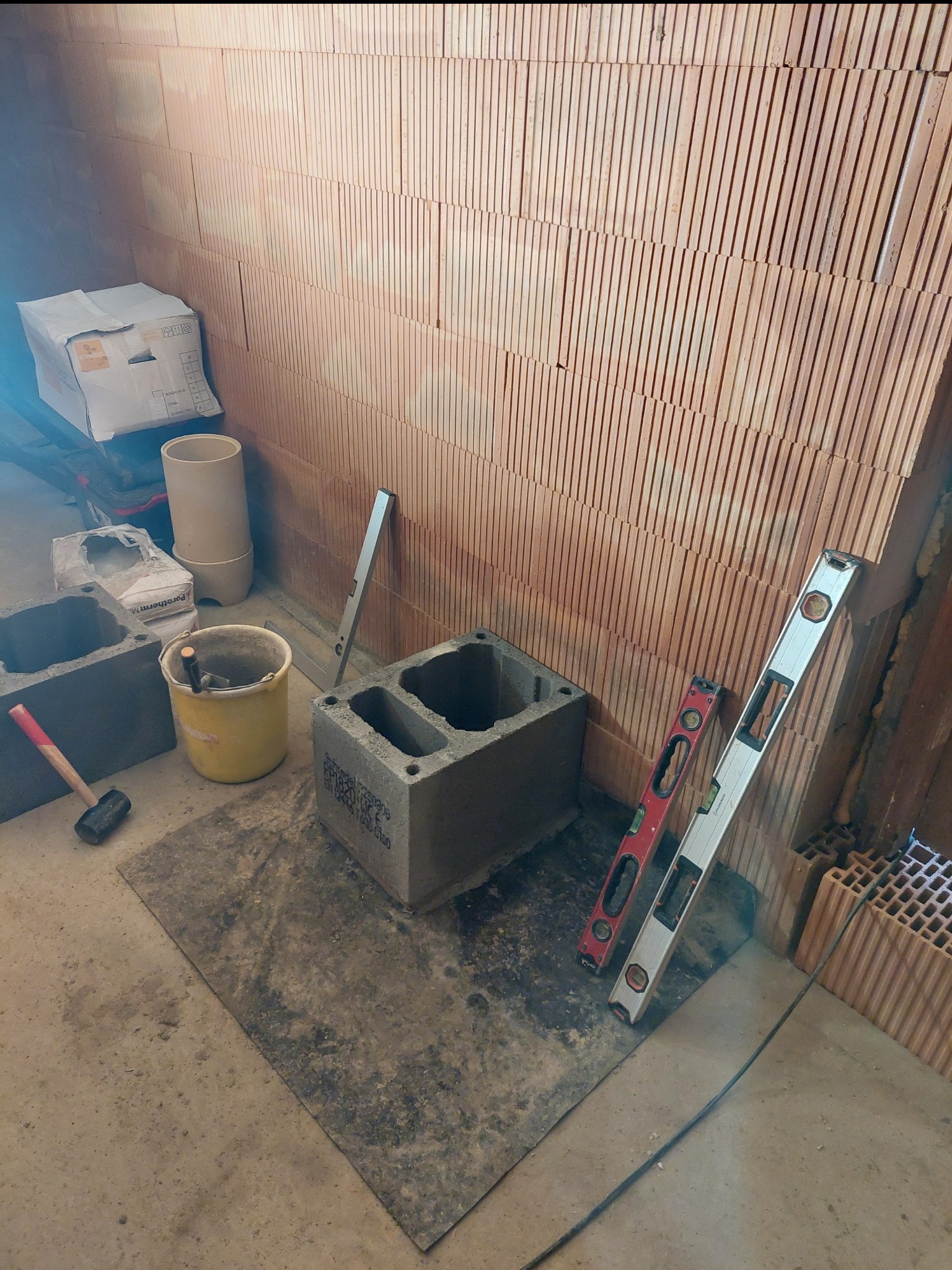 Wnętrze w trakcie budowy: ściana z pustaków ceramicznych, bloczki betonowe, poziomice, wiadro z narzędziami i młotek. Widoczne materiały budowlane.