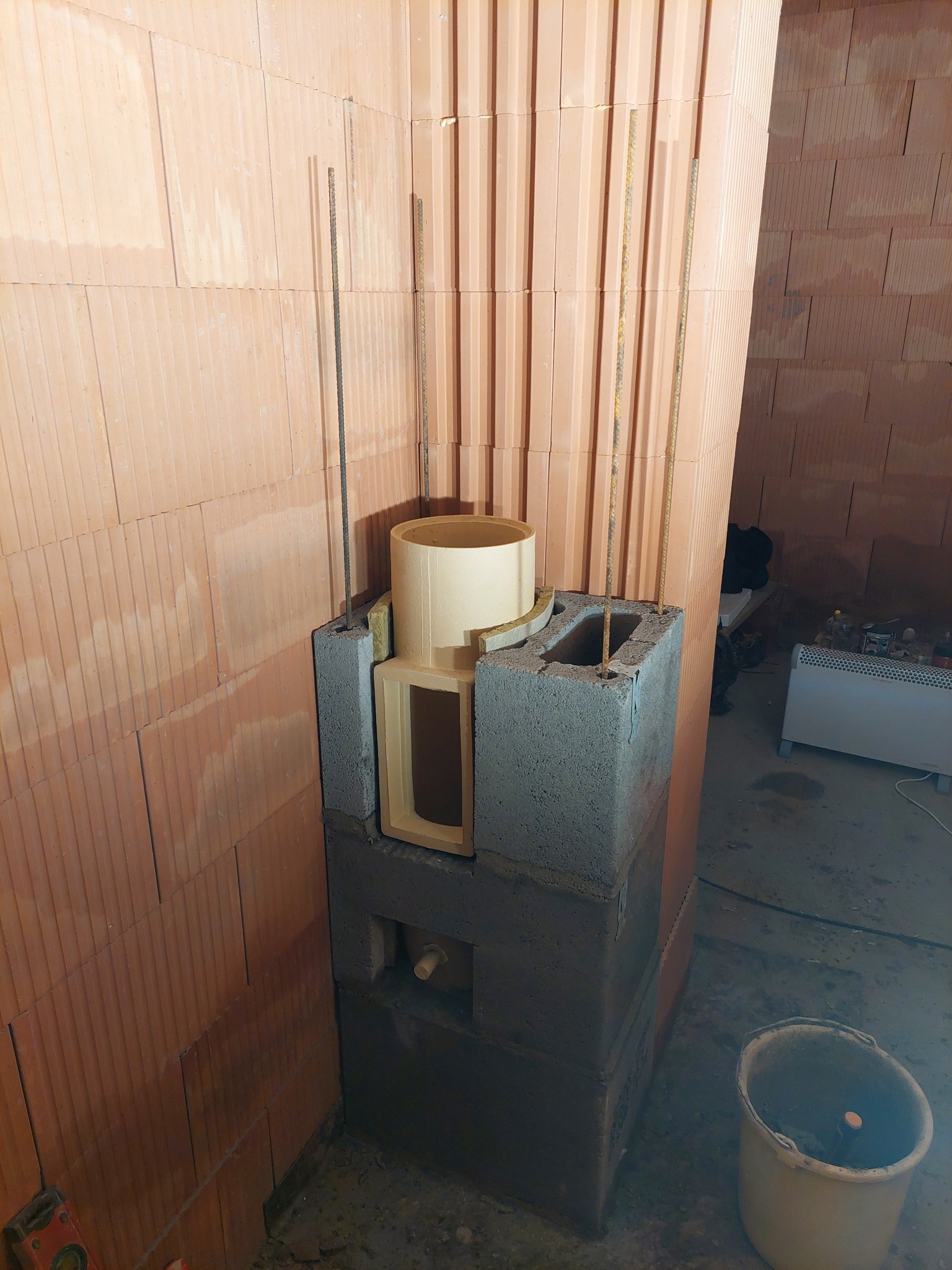 Wznoszenie kominka z szarego bloczka w surowym wnętrzu. Widoczne pręty zbrojeniowe, ceglana ściana i wiadro. Etap budowy kominka w nowym domu.