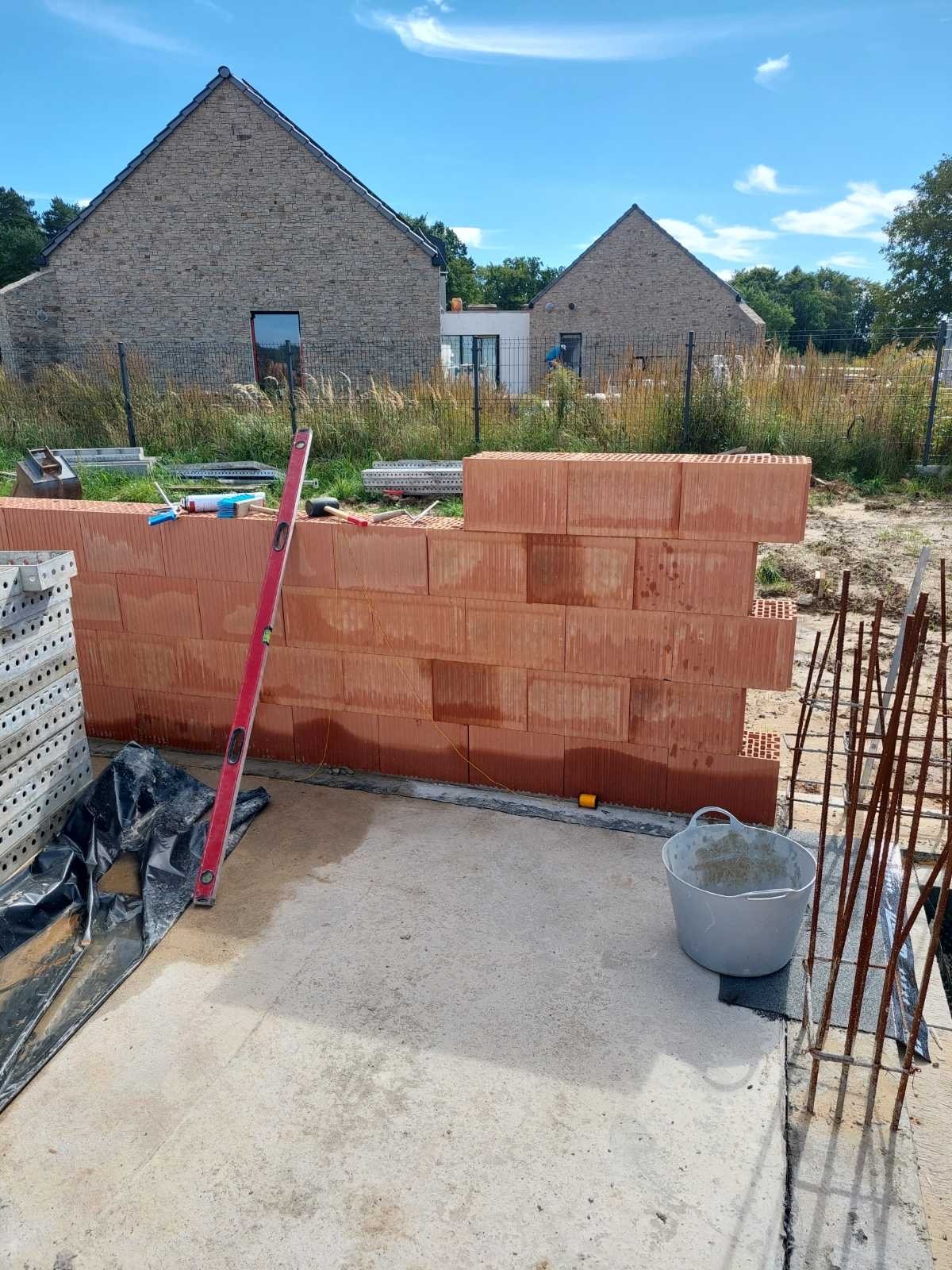 Wznoszenie ściany z czerwonej cegły ceramicznej na fundamencie. Widoczny poziomica, rusztowanie i zbrojenie. W tle domy jednorodzinne i błękitne niebo.