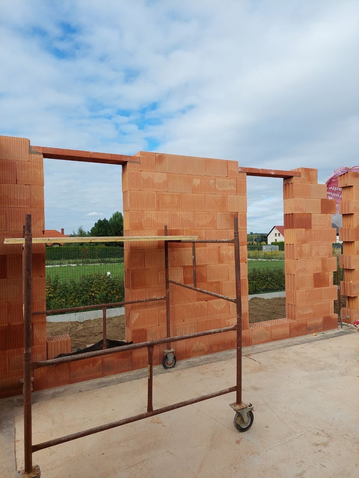 Wznoszenie ścian z czerwonej cegły ceramicznej, widoczne otwory okienne, rusztowanie na kółkach, w tle zieleń i błękitne niebo. Etap budowy domu.