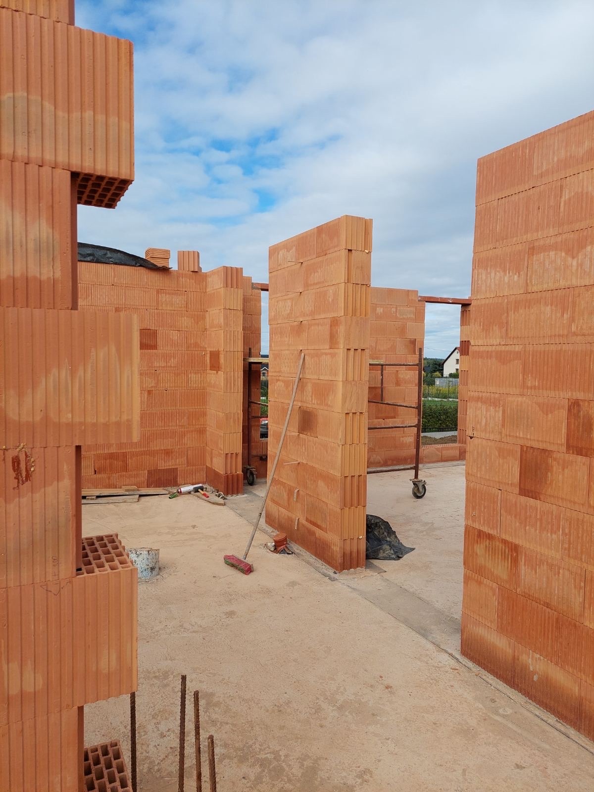 Wznoszenie ścian z czerwonej ceramiki poryzowanej na budowie domu. Widoczne rusztowanie, narzędzia murarskie i fragment nieba. Ściany w trakcie budowy.