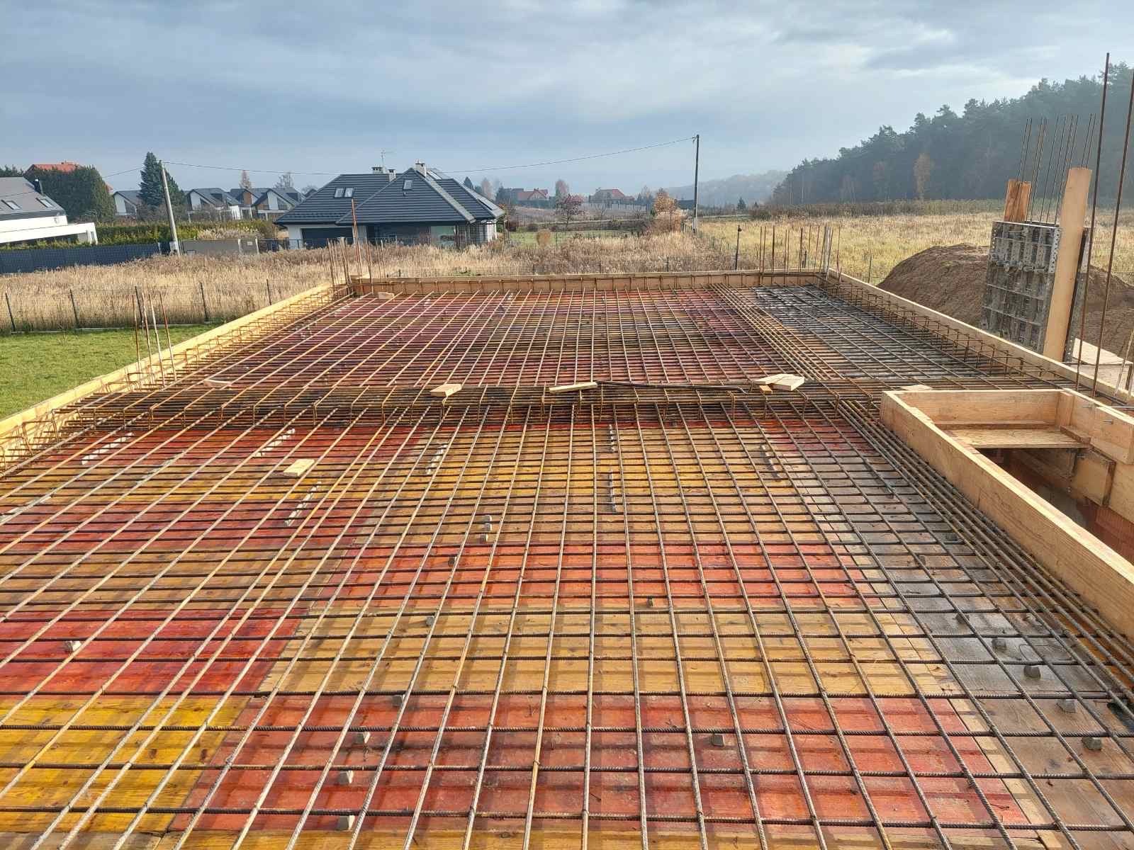 Zbrojenie płyty fundamentowej na deskowaniu, widok z góry. W tle zabudowa jednorodzinna i zalesione wzgórze. Prace zbrojarskie w toku, przygotowanie do zalewania betonem.