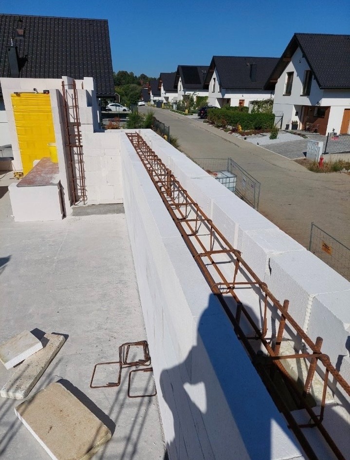 Wznoszenie ścian z betonu komórkowego Ytong z widocznym zbrojeniem wewnątrz, na tle zabudowy jednorodzinnej w słoneczny dzień.