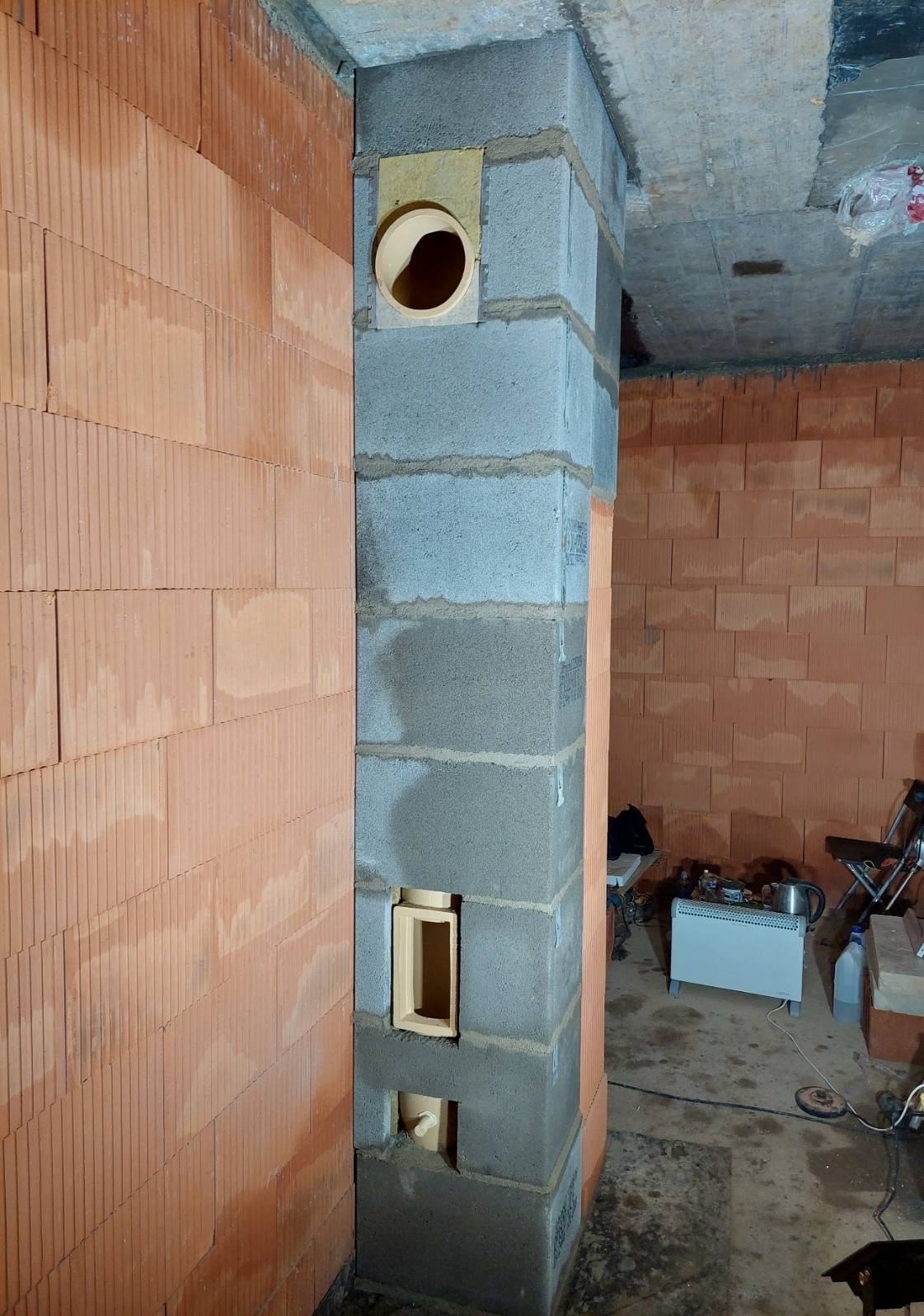 Wznoszenie komina z pustaków szarych, z widocznymi otworami wentylacyjnymi i przyłączeniowymi. Ściana z czerwonej cegły dziurawki w tle. Surowy stan budowy.