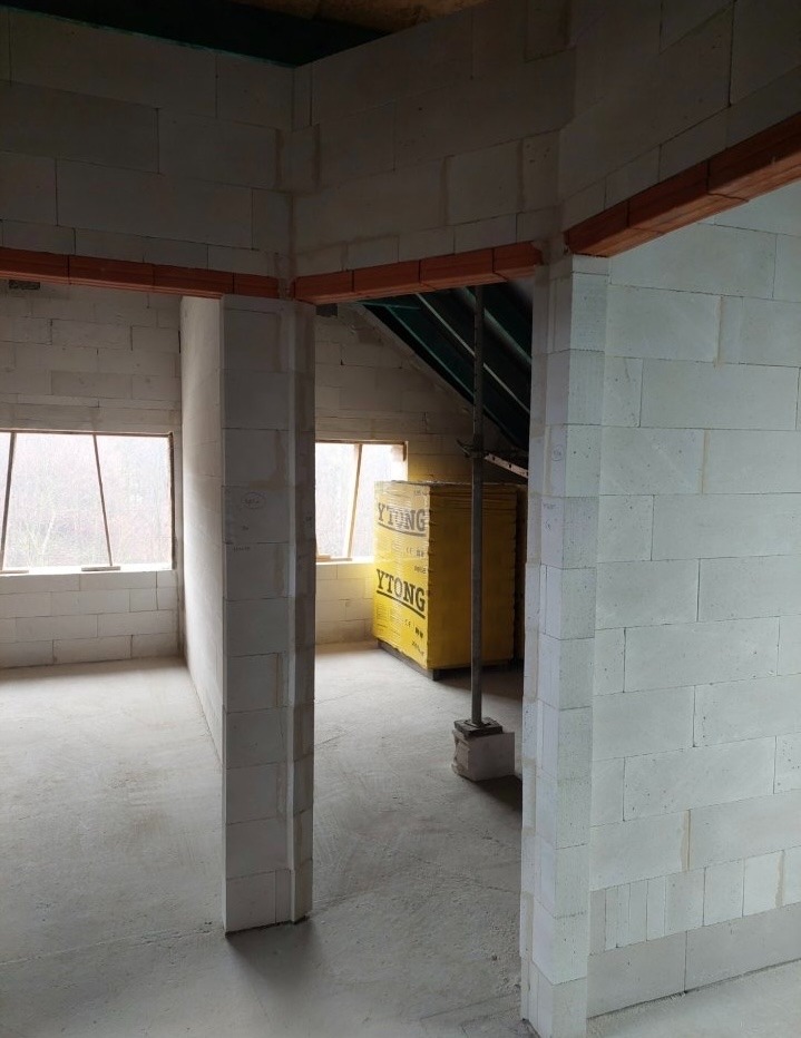 Wnętrze domu w trakcie budowy z białych bloczków Ytong, widoczne okna i drewniane belki stropowe. Na podłodze wylewka, w tle żółte opakowania bloczków.
