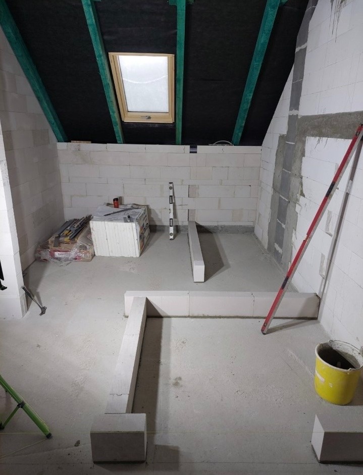 Wnętrze poddasza w trakcie budowy ścian z białych bloczków, widoczne narzędzia, poziomica i wiadro. Ukończona ściana z oknem dachowym. Etap prac murarskich.