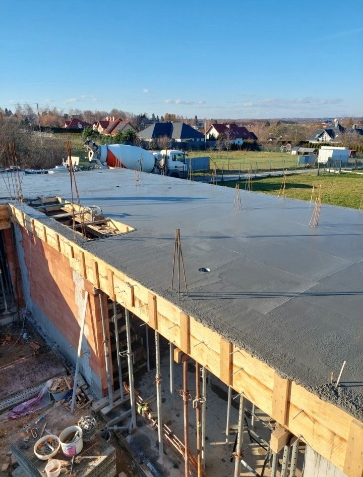 Wylany strop betonowy na budowie domu jednorodzinnego, widoczny mikser do betonu w tle, szalunki i wystające pręty zbrojeniowe. Słoneczny dzień.