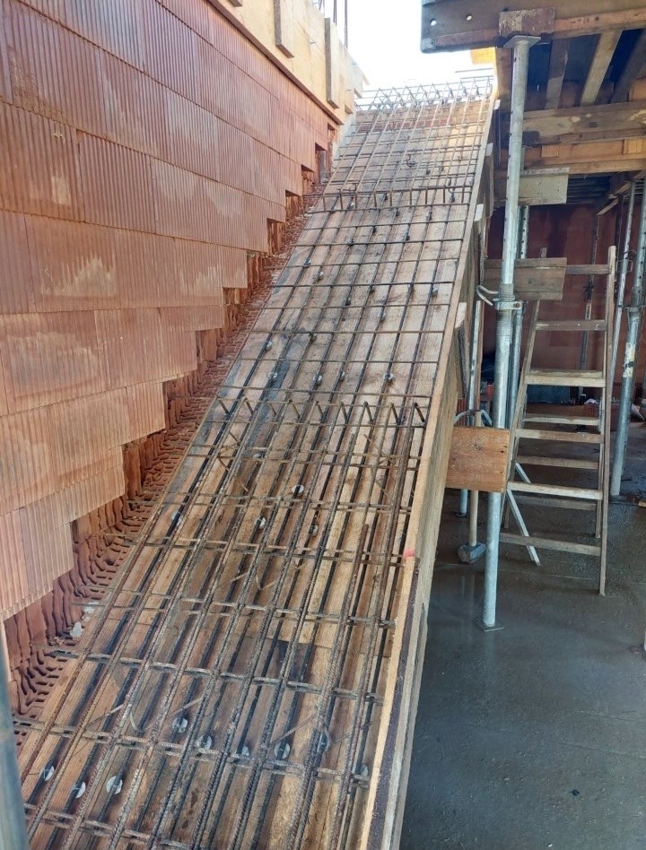 Skośna konstrukcja zbrojeniowa na deskowaniu, widoczna na tle murowanej ściany z pustaków ceramicznych. Elementy drewniane i metalowe podpory.