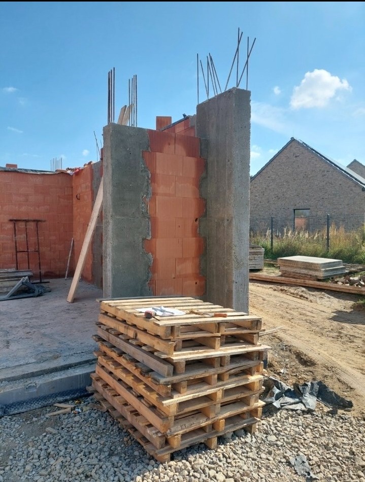 Wznoszenie ścian z pustaków ceramicznych z elementami żelbetowymi. Widoczne wystające pręty zbrojeniowe i paleta z materiałami budowlanymi na placu budowy.