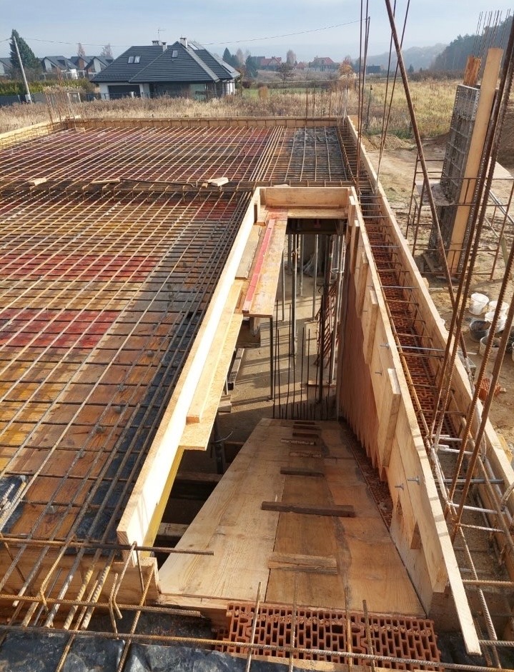 Zbrojenie fundamentów pod dom jednorodzinny z widoczną siatką zbrojeniową, deskowaniem i elementami stalowymi. Prace zbrojarskie w toku, widok z góry.