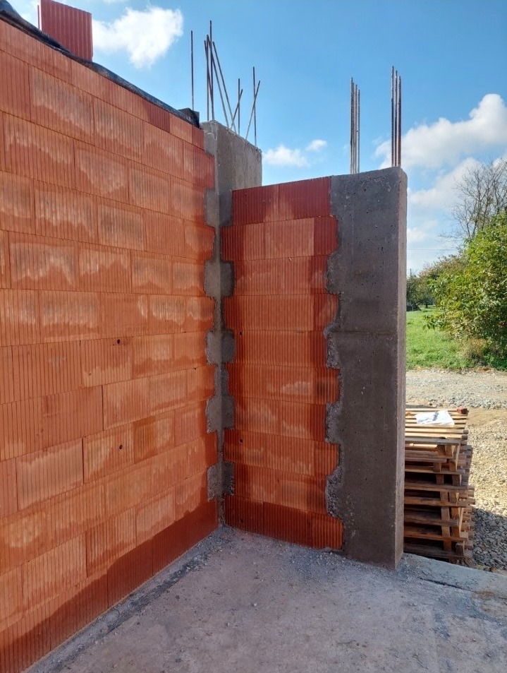 Fragment ściany z czerwonej cegły ceramicznej z betonowym słupem wzmacniającym, widoczne zbrojenie. Budowa w toku, palety w tle.