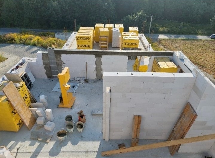 Budowa domu z bloczków Ytong: wznoszone ściany, zapasy materiału na stropie, widoczny podnośnik do bloczków. Ujęcie z góry, w tle zieleń.