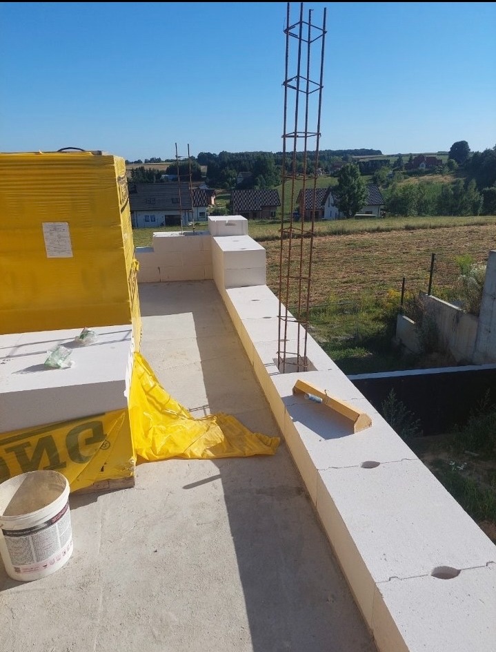 Wznoszenie ścian z bloczków betonu komórkowego Ytong. Widoczna zbrojona kolumna i poziomica. W tle krajobraz z zabudową jednorodzinną.