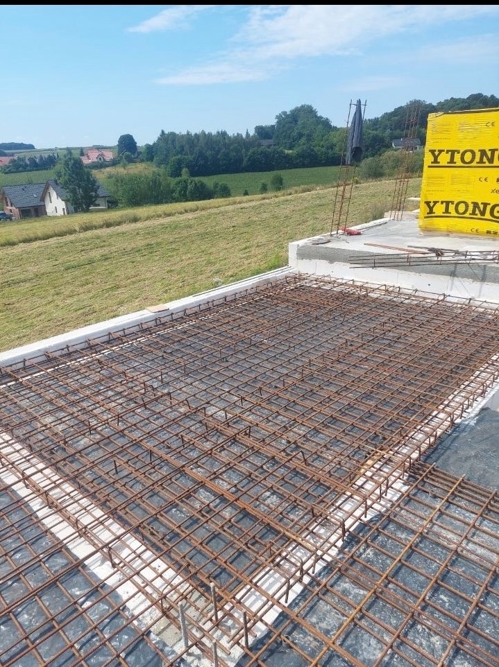 Zbrojenie płyty fundamentowej na budowie domu jednorodzinnego, widok z góry na siatkę z prętów stalowych, w tle zielone pola i zabudowa mieszkalna.