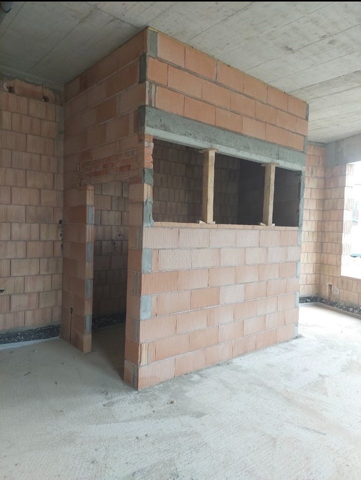 Wnętrze w trakcie budowy z widocznymi ścianami z czerwonej cegły, otworem okiennym z drewnianymi podporami i surowym betonowym sufitem. Widoczny etap prac murarskich.