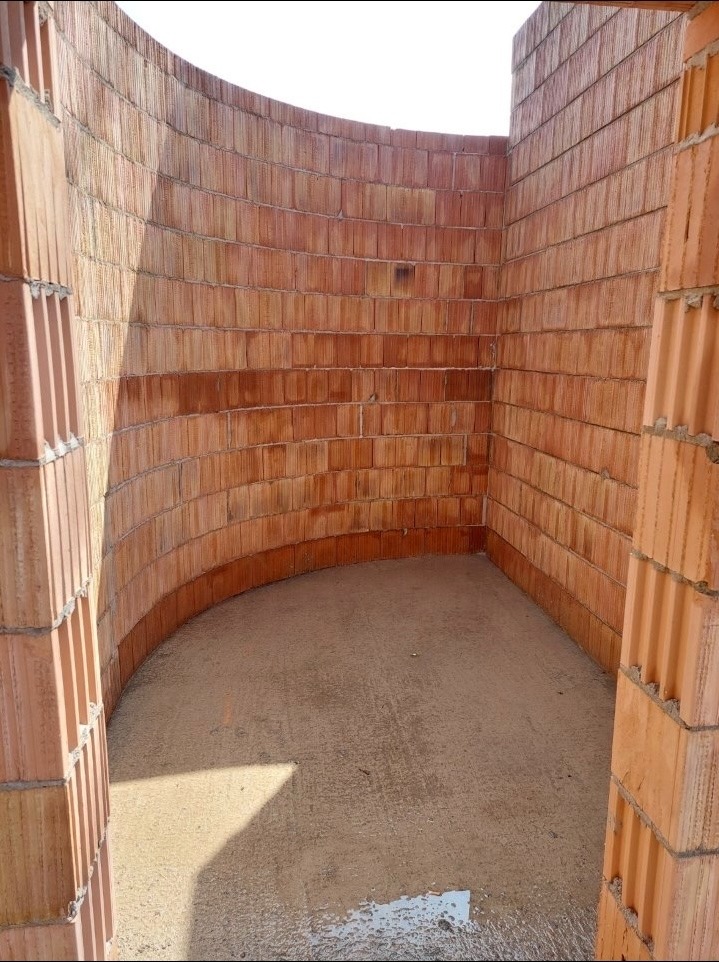 Wnętrze budynku w trakcie budowy z czerwonej cegły, widok na zakrzywioną ścianę i surową podłogę. Fragment ściany po prawej stronie kadru.