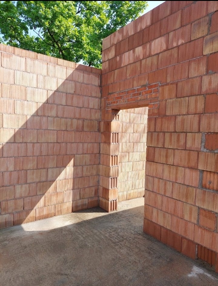 Wnętrze budynku w trakcie budowy z czerwonej cegły, widoczne fragmenty ścian i otwór drzwiowy. Na ścianie cień, w tle zieleń drzew.