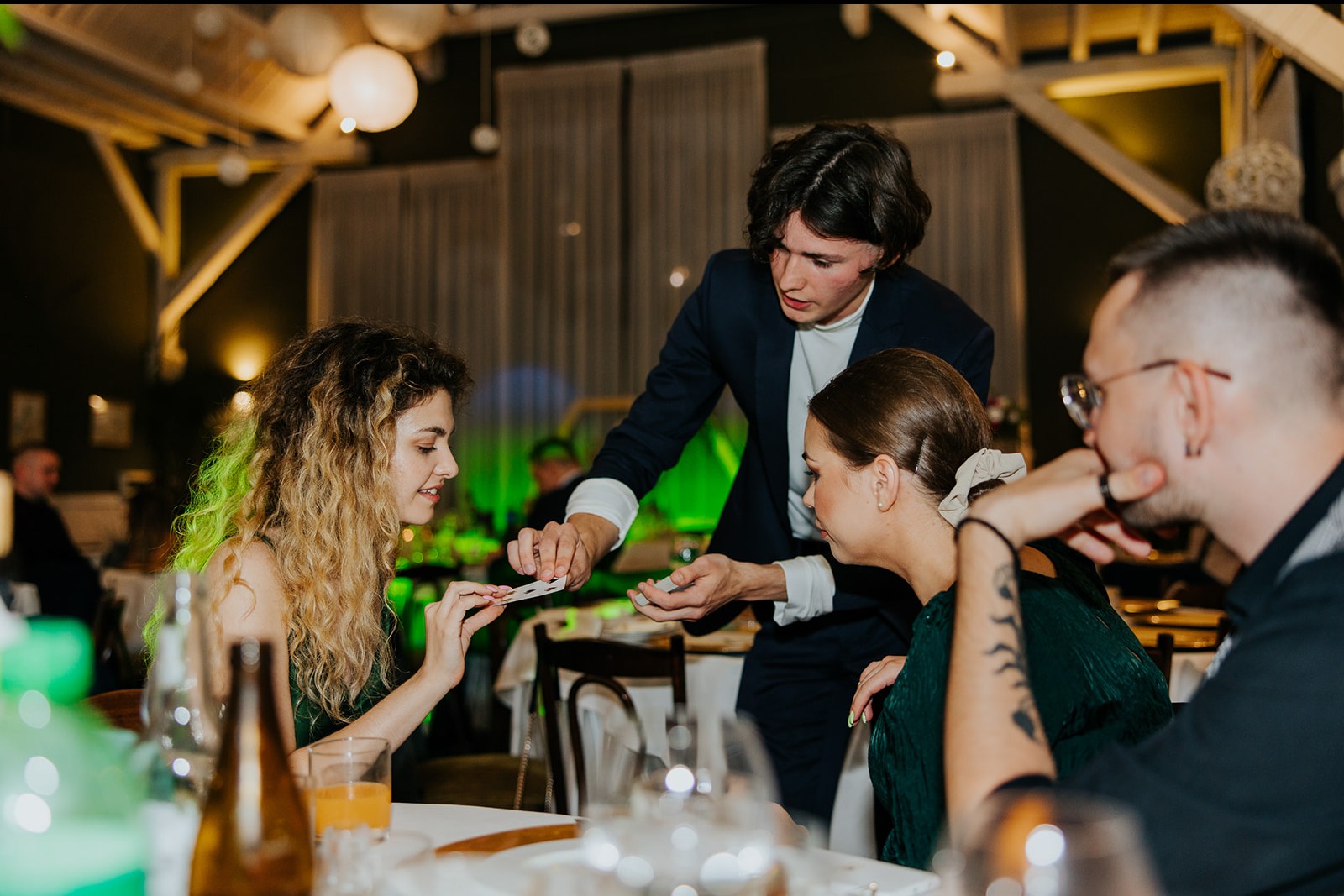 Iluzjonista w granatowym garniturze prezentuje trik karciany grupie gości siedzących przy stole podczas pokazu close-up w oświetlonym wnętrzu.