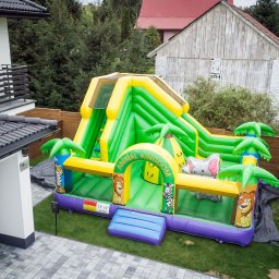 Plenerowo.com Atrakcje & Gastronomia - Kolorowy, dmuchany zamek z motywem zwierząt, ustawiony na trawniku obok domu. Widoczny fragment elewacji budynku i zieleń w tle. Zamek ma zjeżdżalnię i elementy dekoracyjne.